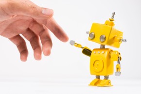 Human hand touches a tiny yellow robot's hand